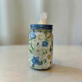 A mason jar tissue holder with a blue lid and a floral paper wrap featuring blue flowers and white daisies on a light green plaid background, with white tissues visible at the top, set on a light wooden surface against a soft beige background.